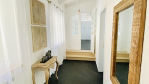 Een gang met een houten vloer, witte wanden en een grote spiegel met ingewikkelde versieringen. Twee ingelijste foto's hangen aan de muur en er staat een klein tafeltje met decoratieve objecten.