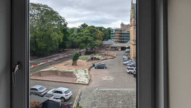Vista a través de una ventana que muestra un estacionamiento con coches, un edificio con andamios y árboles.