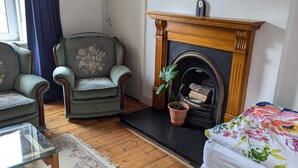 Una habitación con chimenea, dos sillones verdes, una mesa de centro de vidrio, una cama con ropa de cama floral y una planta en maceta.