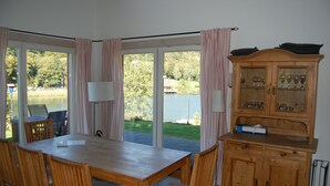 Et spiseområde med et bord og stole, et træskab med glasdøre, og udsigt til en sø gennem store vinduer.