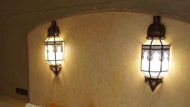 A bed with white bedding and two orange pillows, two wall-mounted light fixtures, and a textured wall. - Dar Lkhalifa Merzouga (Taouz)