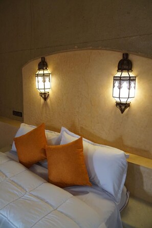 A bed with white bedding and two orange pillows, flanked by two wall-mounted light fixtures. - Dar Lkhalifa Merzouga (Taouz)