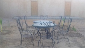 A round metal table with four chairs, set on a concrete floor in a courtyard with a plain wall and two wooden doors in the background. - Dar Lkhalifa Merzouga (Taouz)