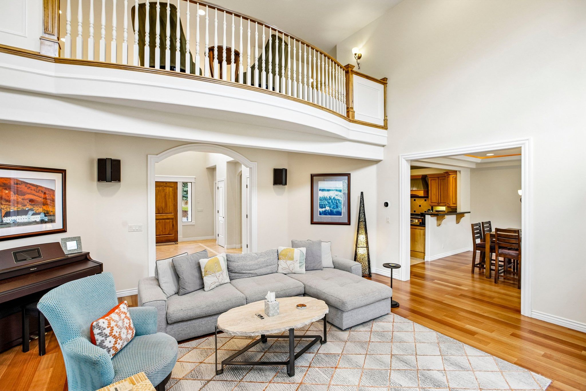 A spacious living room with a sectional sofa, a round coffee table, and a piano. A balcony with a railing is visible above.