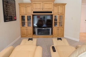 Una sala de estar con un televisor de pantalla plana grande, armarios de madera y dos sofás color beige con controles remotos.