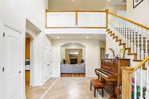 A spacious foyer with a grand staircase, a piano, and a kitchen area in the background.