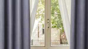 A window with white curtains, a radiator below, and a view of a building and trees outside. - Magik Rooms (Rome)