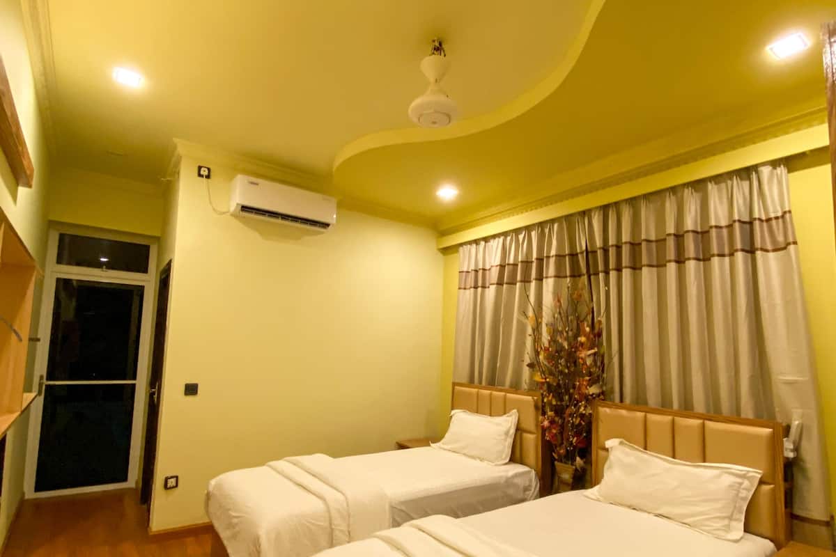 A hotel room with two beds, wooden flooring, a ceiling fan, an air conditioning unit, and a window with curtains.