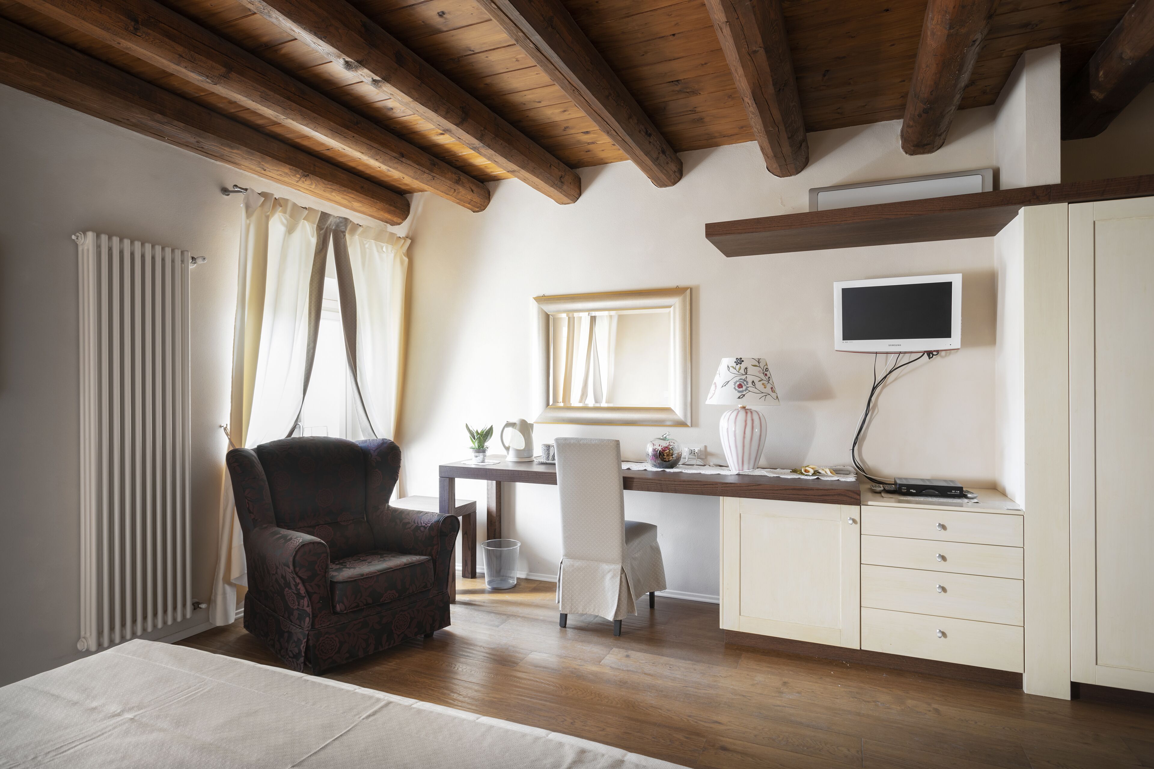 A bedroom with a wooden ceiling, a bed, a desk with a chair, a television, and a radiator.