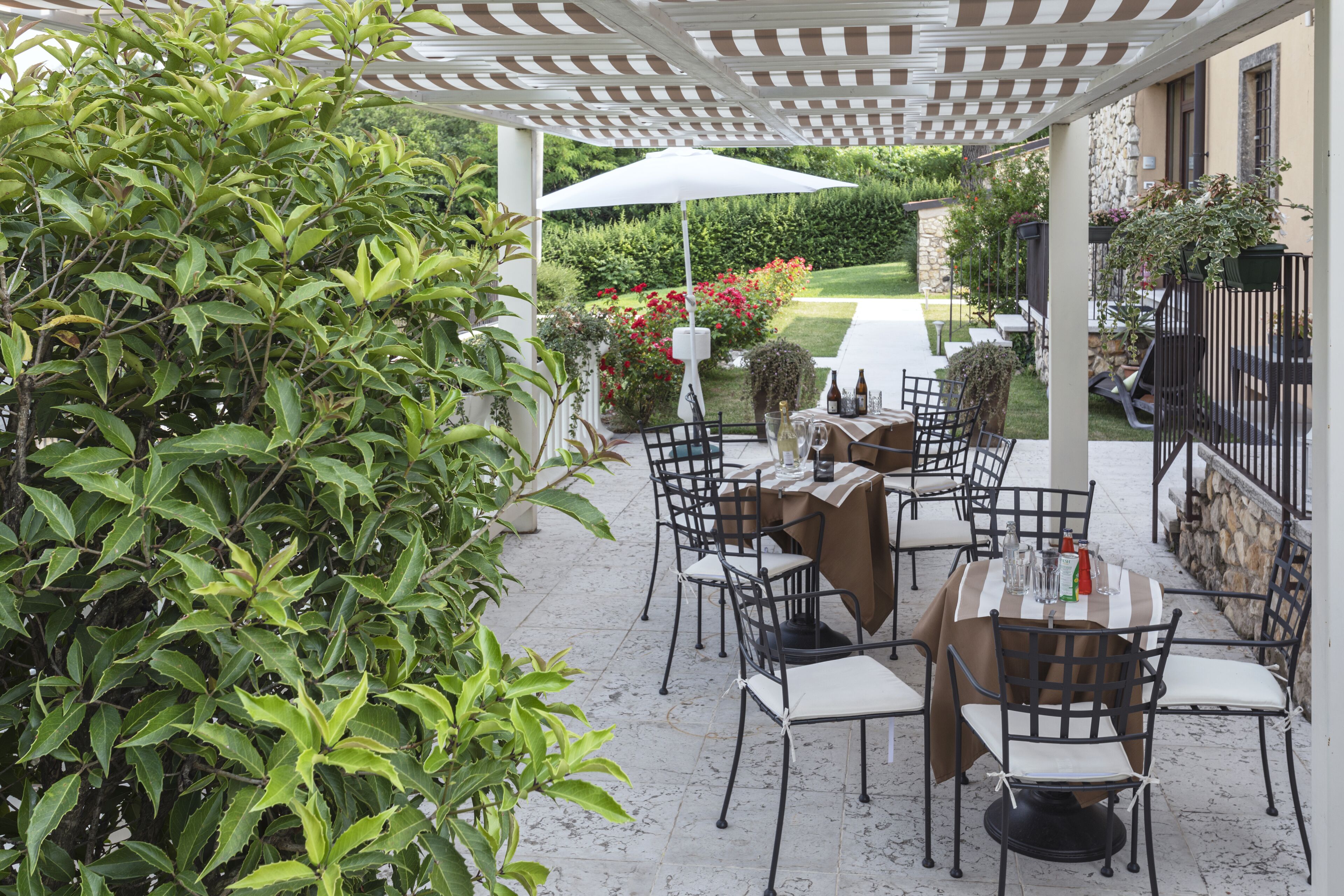 An outdoor patio with tables and chairs, surrounded by greenery and a white pergola.