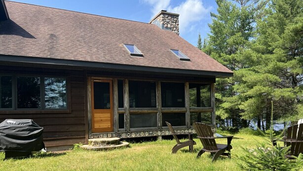 A wooden cabin with a sloped roof, large windows, and a porch with two Adirondack chairs. - Piper`s Point on Lake Julia (Hiles)