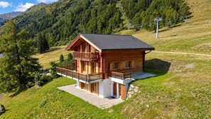 Eine Holzhütte mit Balkon, eingebettet in eine Berglandschaft, im Hintergrund eine Seilbahn.