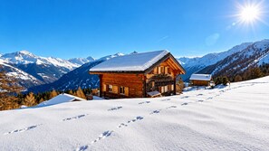Eine Holzhütte mit schneebedecktem Dach, umgeben von Bergen und blauem Himmel.