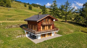 Un chalet en bois doté d’un balcon, situé sur une colline verdoyante, avec des arbres en arrière-plan.