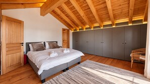 Ein Schlafzimmer mit einem Bett, einem grauen Kleiderschrank, Holzbalken an der Decke und einem Korbstuhl.