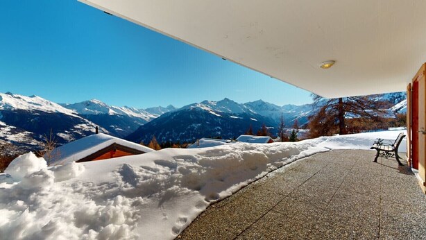 Un sentier enneigé mène à un chalet en bois doté d’un banc, le tout étant entouré de montagnes et surplombé par un ciel dégagé.