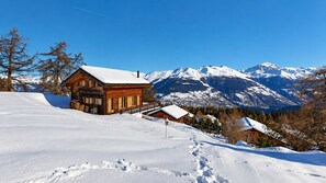 Un chalet en bois, dont le toit est recouvert de neige, entouré de montagnes et d’arbres également couverts de neige.