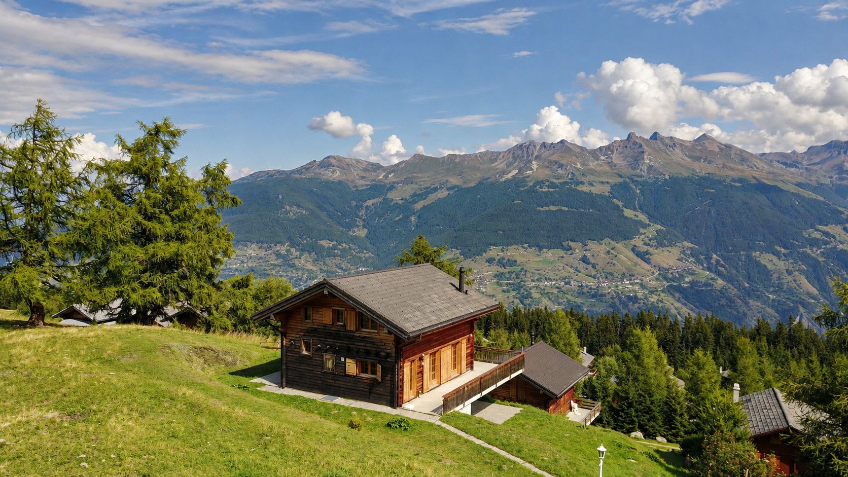A wooden cabin with a balcony, surrounded by lush greenery and mountains in the background.