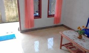 A room with a tiled floor, a wooden table with a vase of flowers, and two windows with pink curtains. - A small house in Sorong City (Sorong)