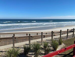 A wooden boardwalk leading to a beach with waves, a clear sky, and a sandy area with some grass.