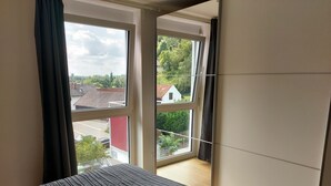 Un dormitorio con cama, ventana con cortinas y vista a la calle y vegetación.