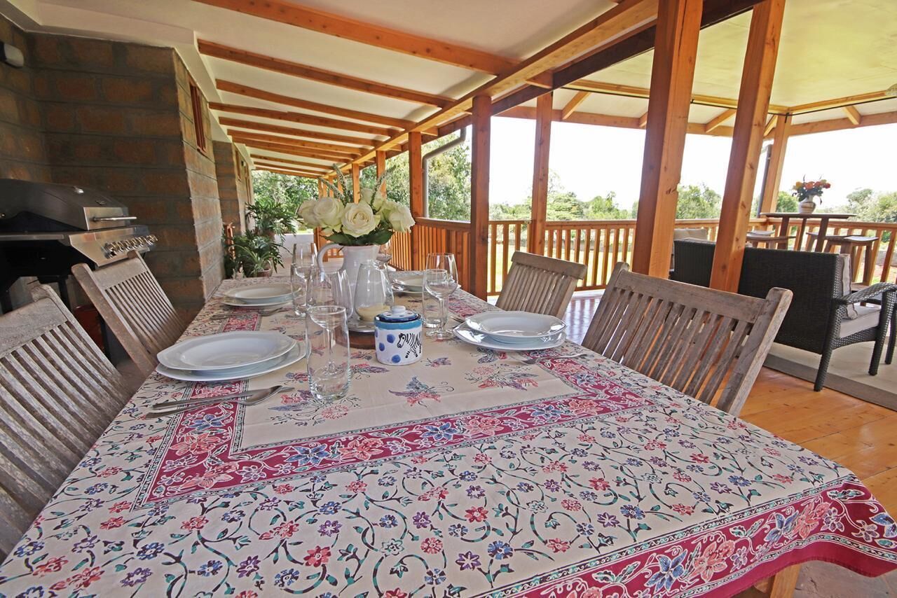Un área de comedor techada con una mesa dispuesta para cuatro personas, rodeada de sillas de madera, y una parrilla a la izquierda.