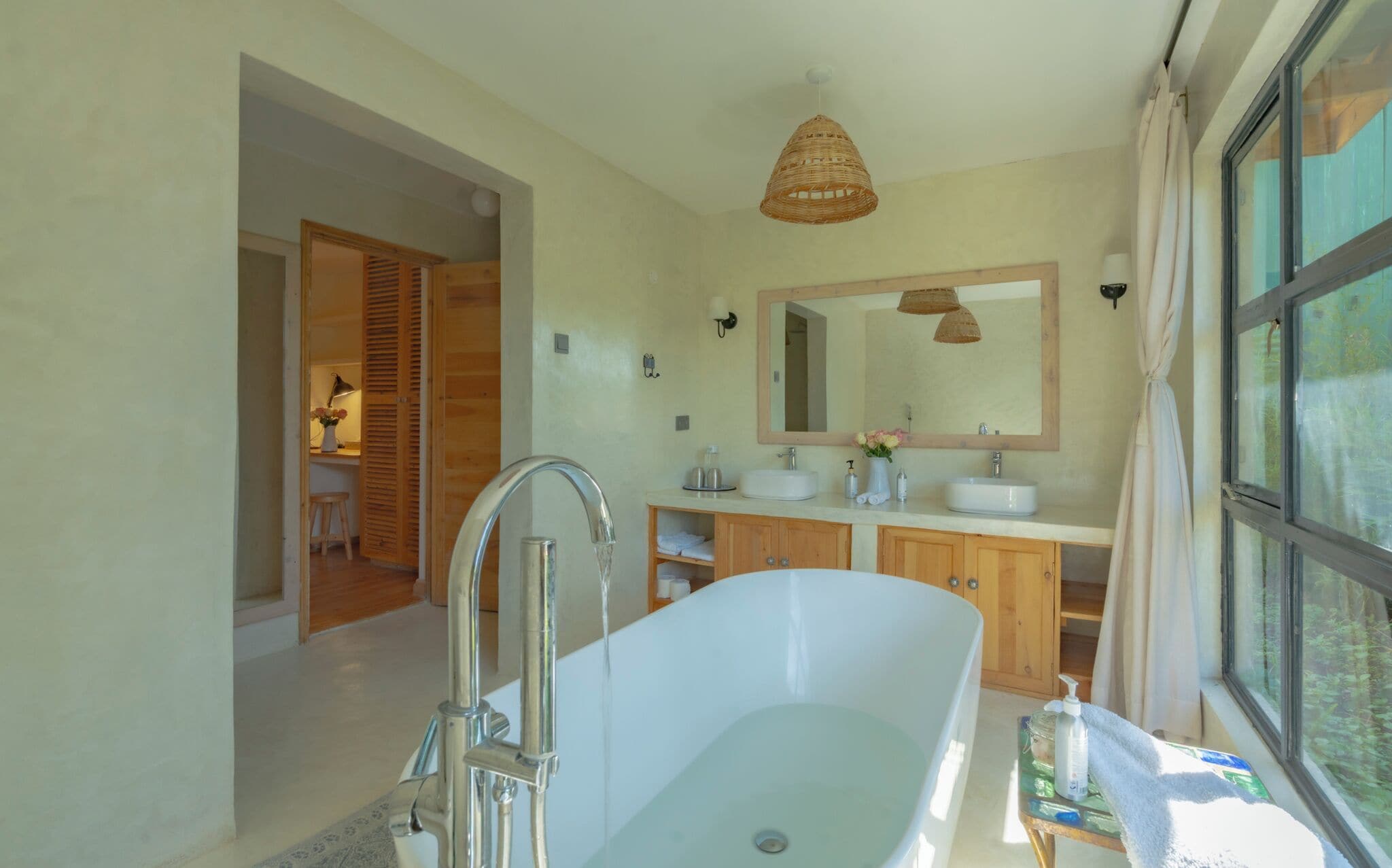 A modern bathroom with a freestanding bathtub, wooden cabinets, a large mirror, and a window providing a view of greenery.