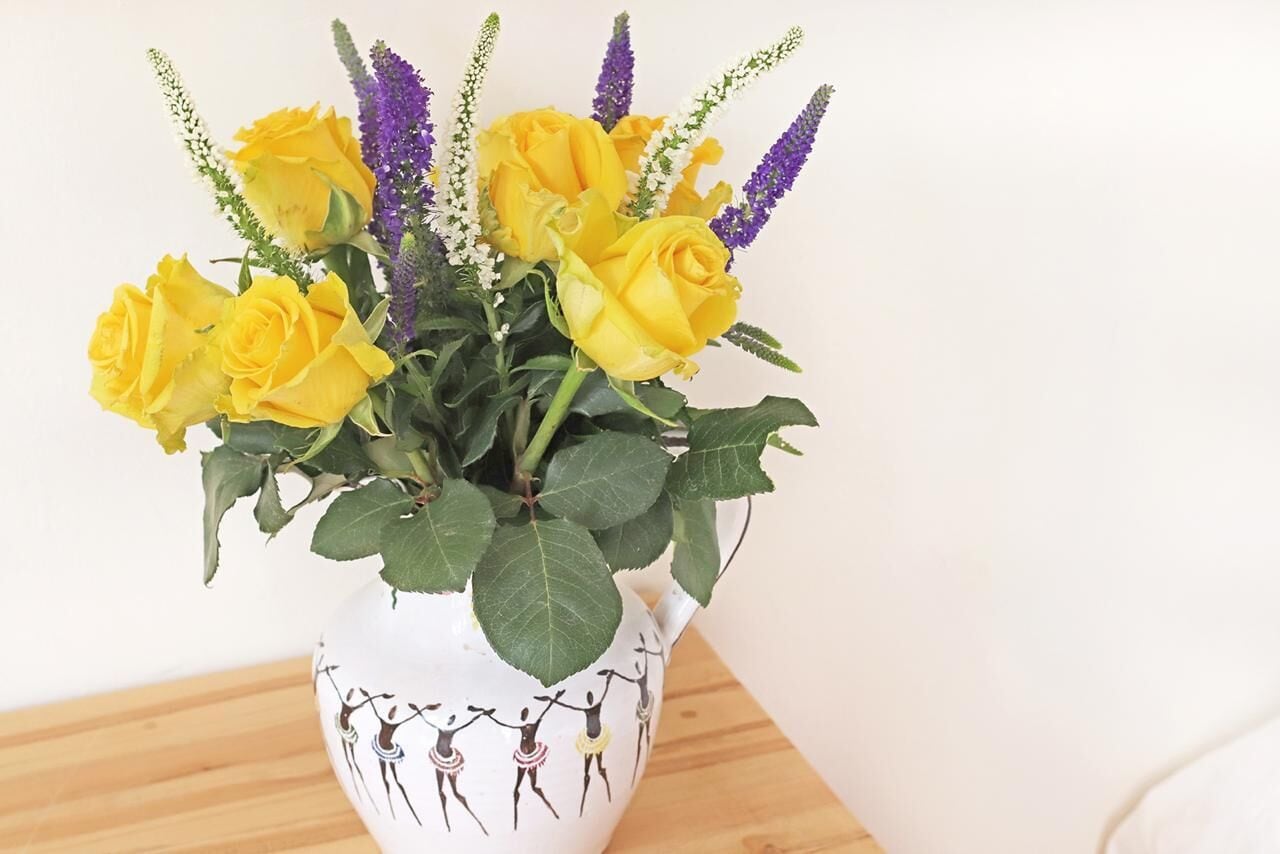 Um vaso branco com arranjo floral de rosas amarelas e flores roxas.