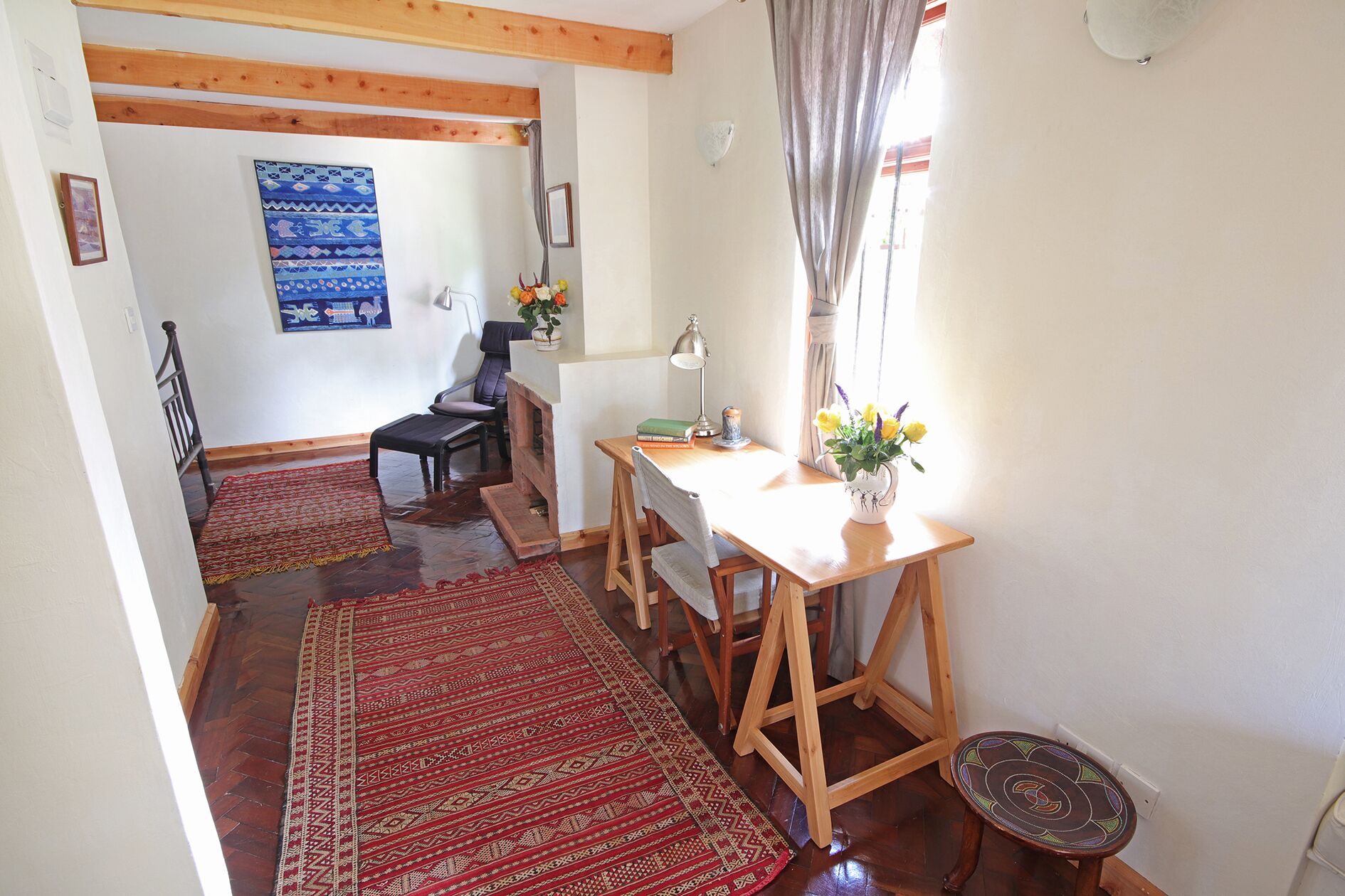 A cozy room with a wooden table, a chair, a small stool, a rug, and a window with curtains.