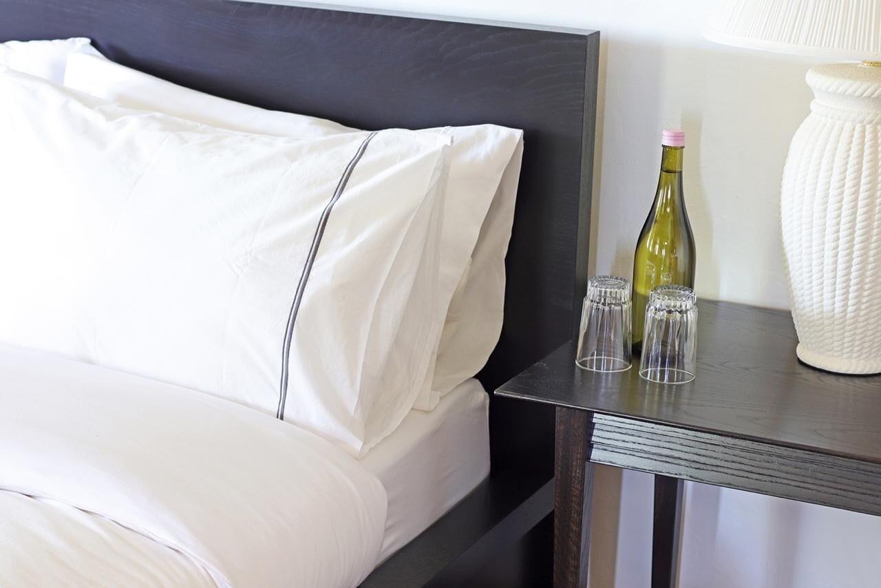 Un lit bien fait, avec du linge de lit blanc, une tête de lit sombre et une petite table à côté, sur laquelle se trouvent une bouteille et des verres.