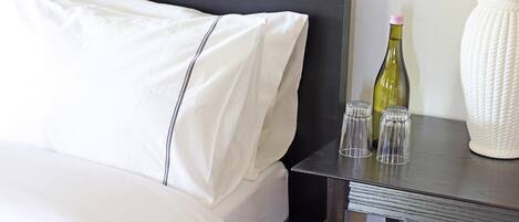 Una cama bien hecha con sábanas blancas, un cabecero oscuro y una mesita de noche con una botella y vasos.