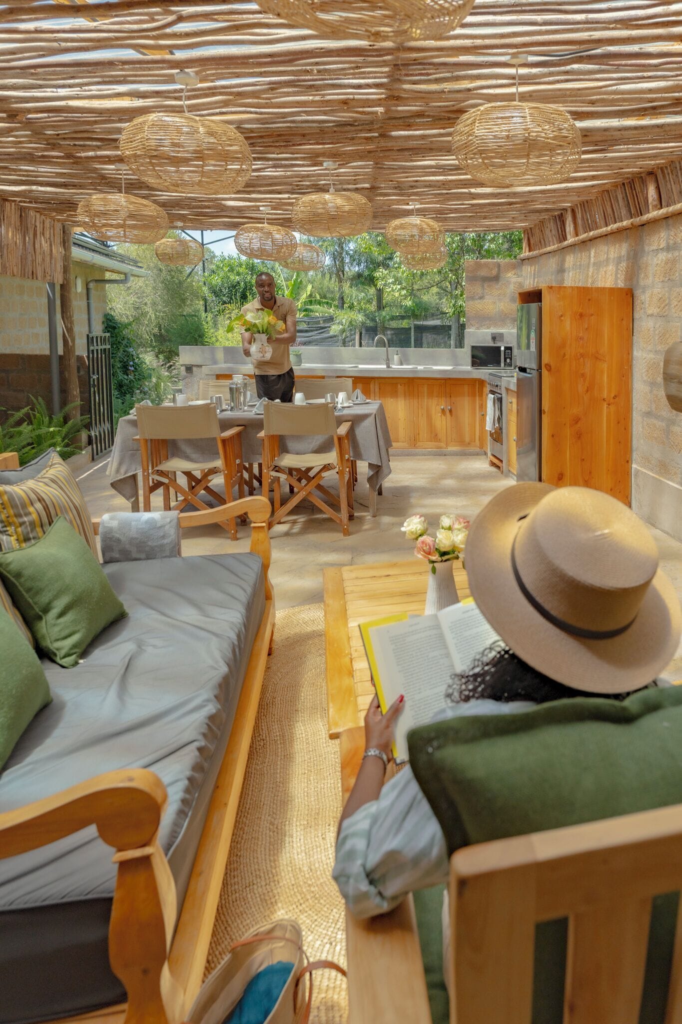 Un área de comedor al aire libre y acogedora con muebles de mimbre, una mesa de madera y una persona leyendo un libro.