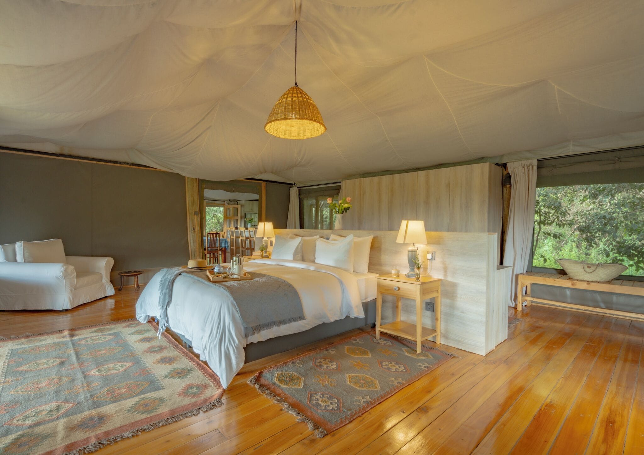 A spacious bedroom with a large bed, bedside tables, a hanging light fixture, and a view of greenery outside.