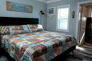 A bedroom with a bed, a wall-mounted air conditioner, a window with blinds, and a door.