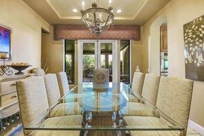 A dining area with a glass-top table surrounded by upholstered chairs, a chandelier, and a view of the outdoors. - Tuscany: Pool, Spa, Fire Pit, Ping Pong Games (Indio)