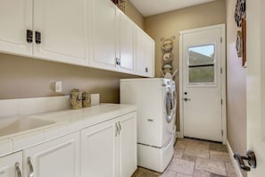 A compact kitchen with white cabinets, a built-in washer and dryer, and a small window. - Tuscany: Pool, Spa, Fire Pit, Ping Pong Games (Indio)