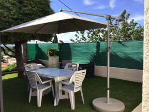 Eine Außenpatio mit Tisch und Stühlen unter einem großen Sonnenschirm, umgeben von grünem Kunstgrün und einem grünen Zaun.