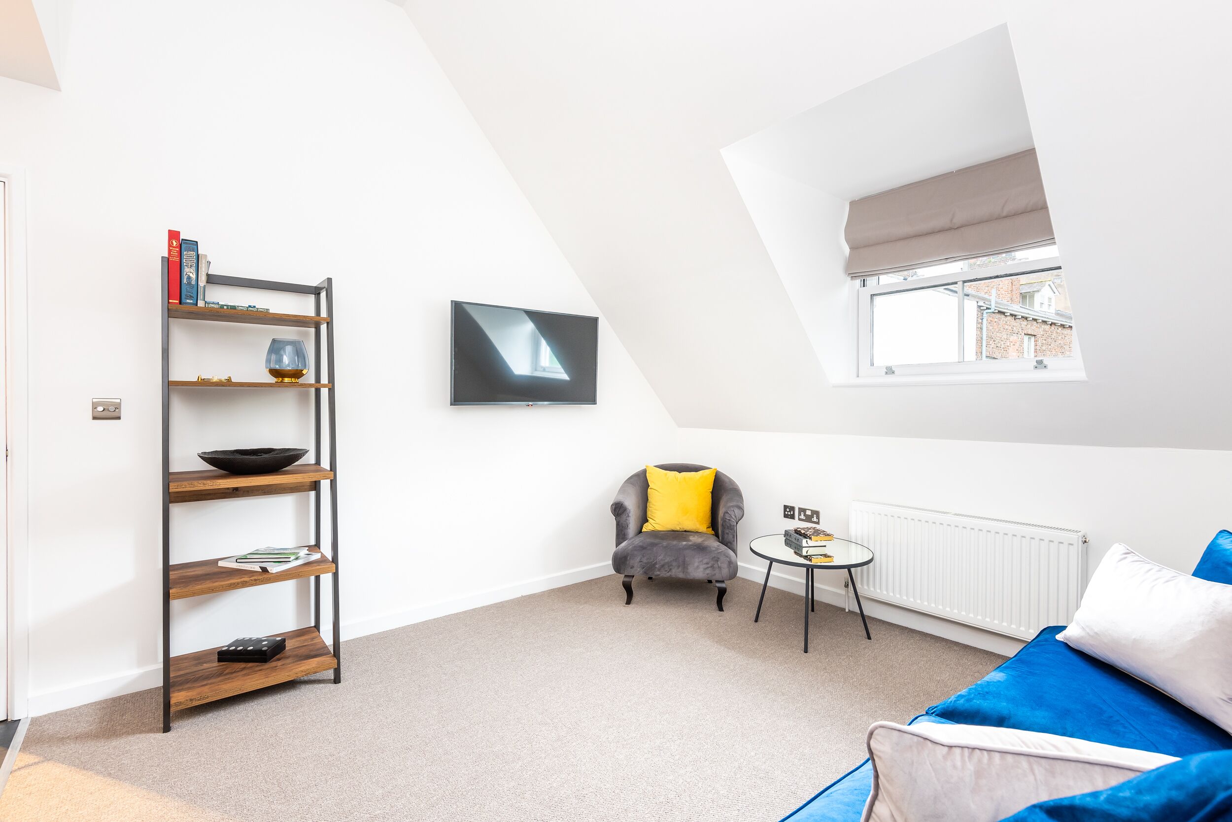 Un salon moderne avec un lit en mezzanine, un fauteuil gris agrémenté d’un coussin jaune, un canapé bleu, une petite table ronde et une bibliothèque adossée au mur.