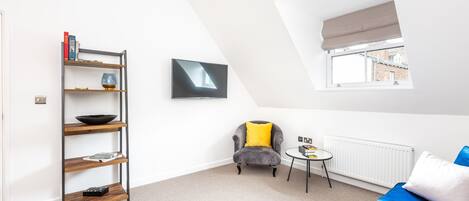 Un salon moderne avec un lit en mezzanine, un fauteuil gris agrémenté d’un coussin jaune, un canapé bleu, une petite table ronde et une bibliothèque adossée au mur.