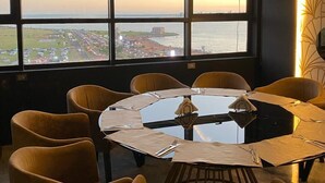 A dining area with a round table and chairs, large windows with a view of a cityscape and water, and pendant lighting. - Sun Hotel (Encarnacion)