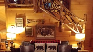 Una sala de estar acogedora con un sofá de cuero oscuro, dos lámparas, un cojín con temática de oso, una escalera de madera y una alfombra con huellas de animales.