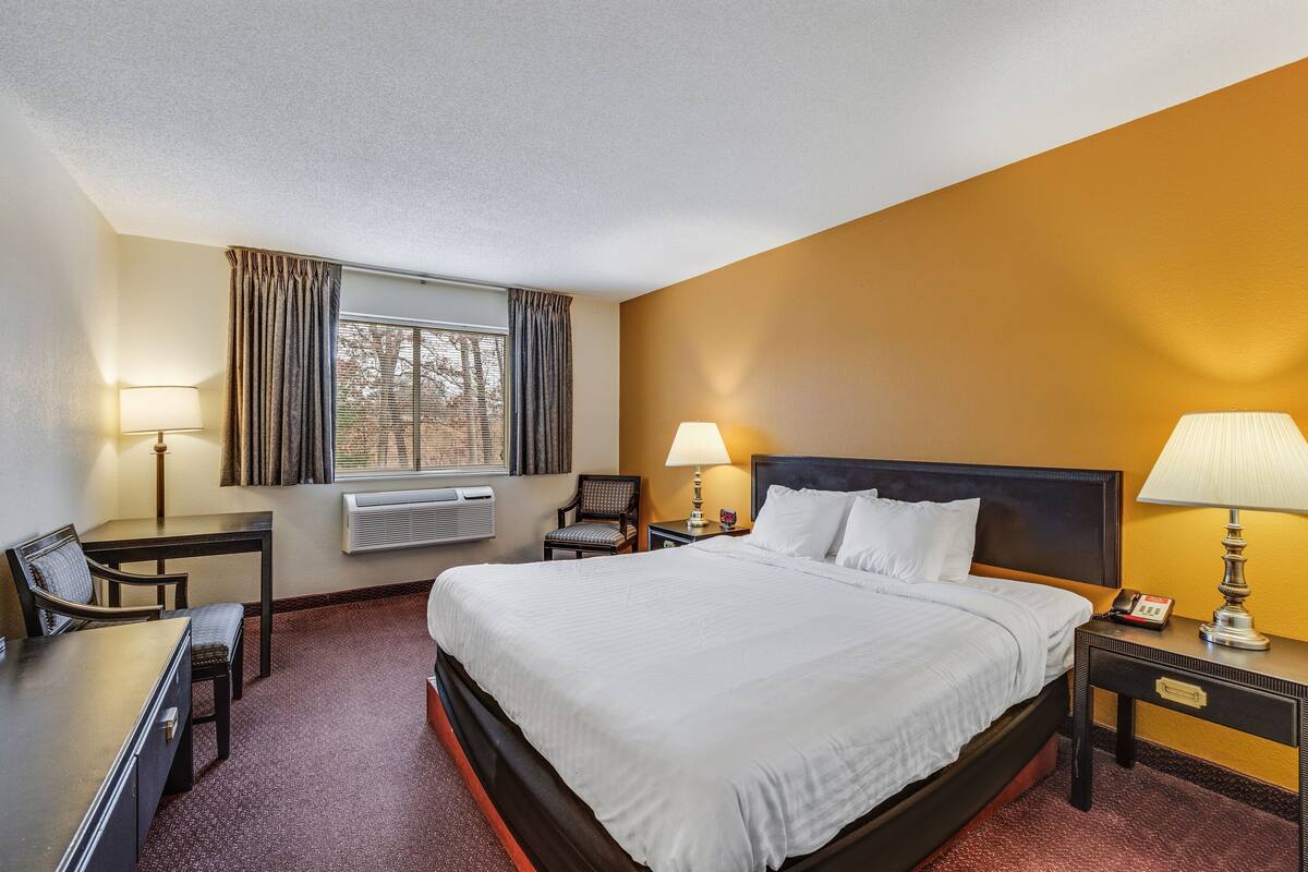 A hotel room with a bed, desk, chair, lamp, and window.