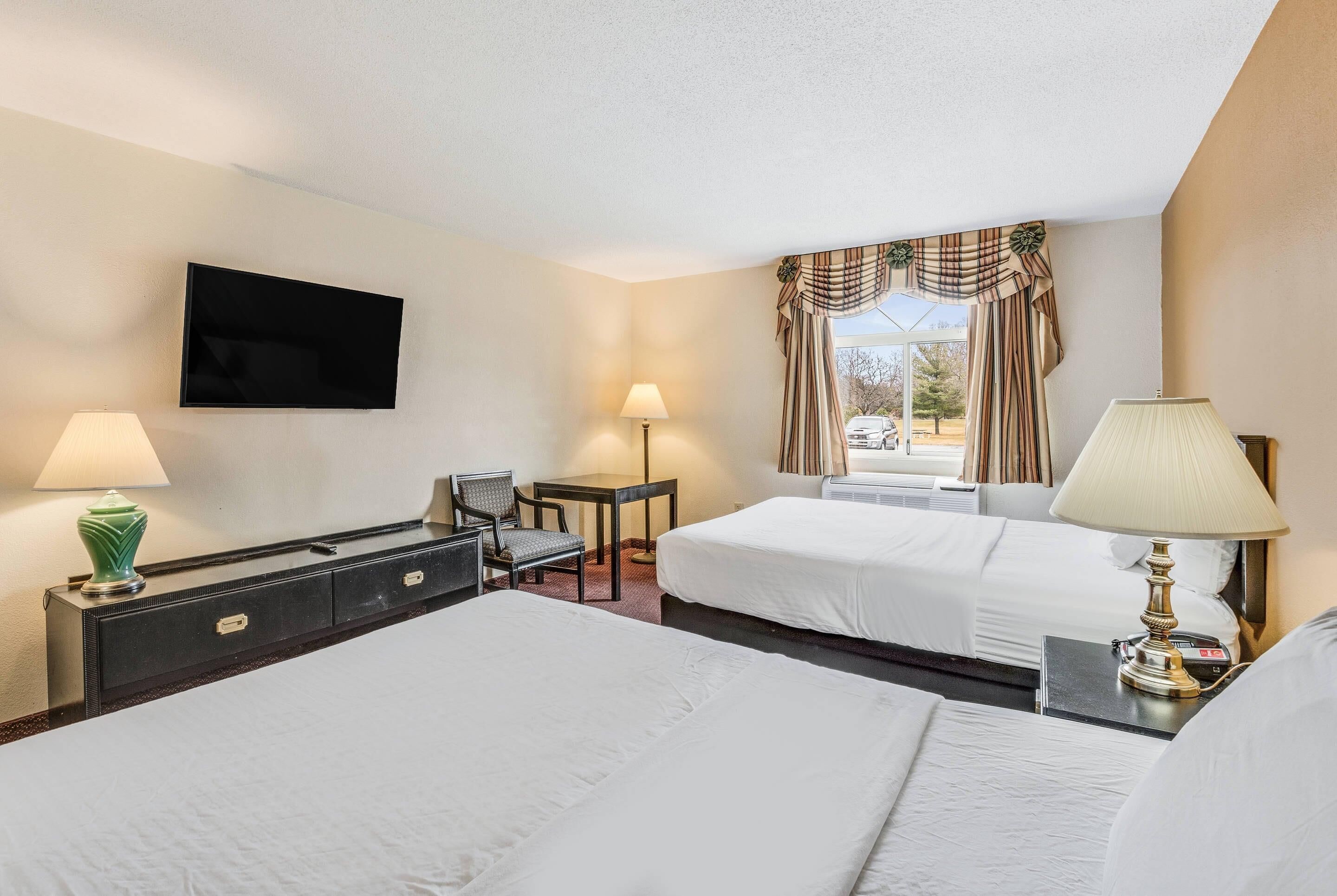 A hotel room with two beds, a flat-screen TV mounted on the wall, a desk with a chair, and two lamps.