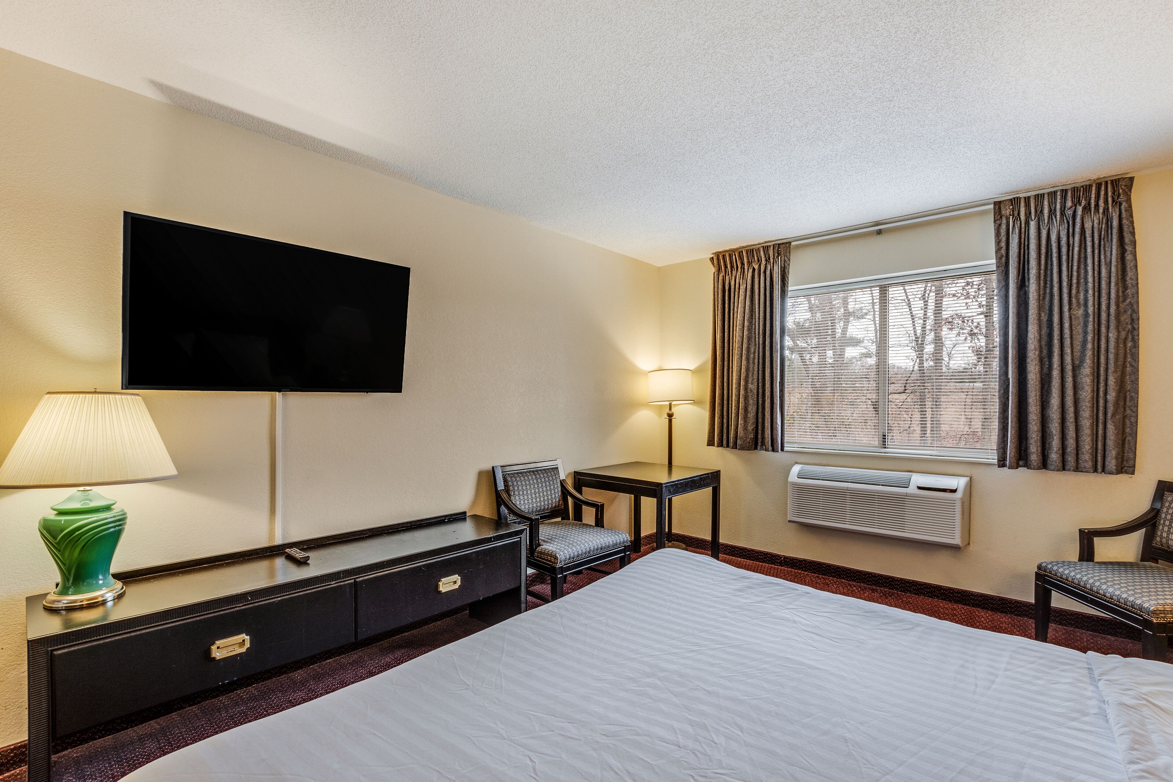 Habitación de hotel con una cama grande, un televisor de pantalla plana en la pared, un escritorio con lámpara, una silla y una ventana con cortinas.