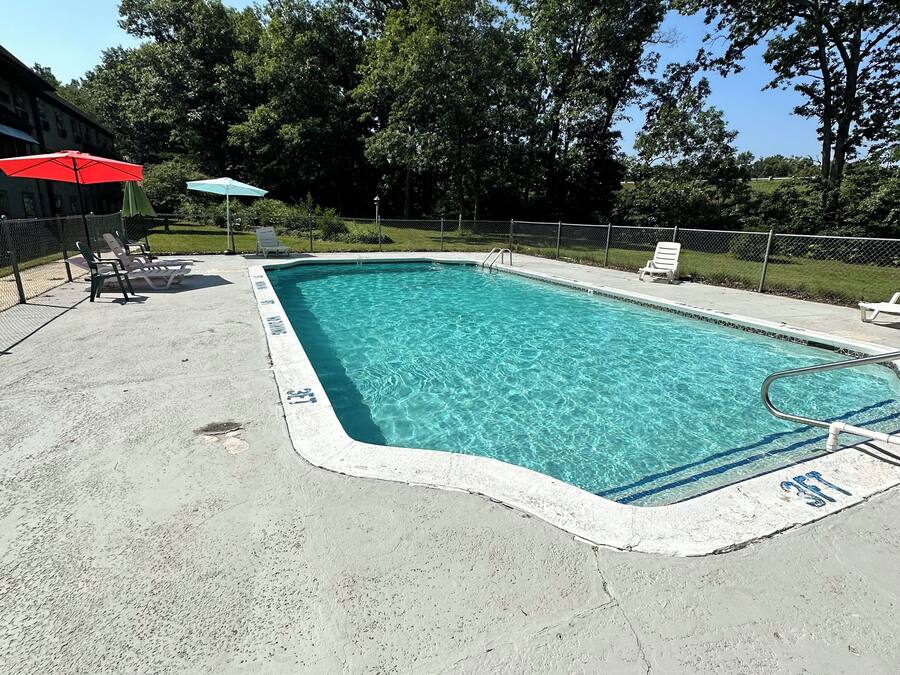 Uma piscina retangular com água azul cristalina, rodeada por um deck de concreto. Há espreguiçadeiras e guarda-sóis à beira da piscina.