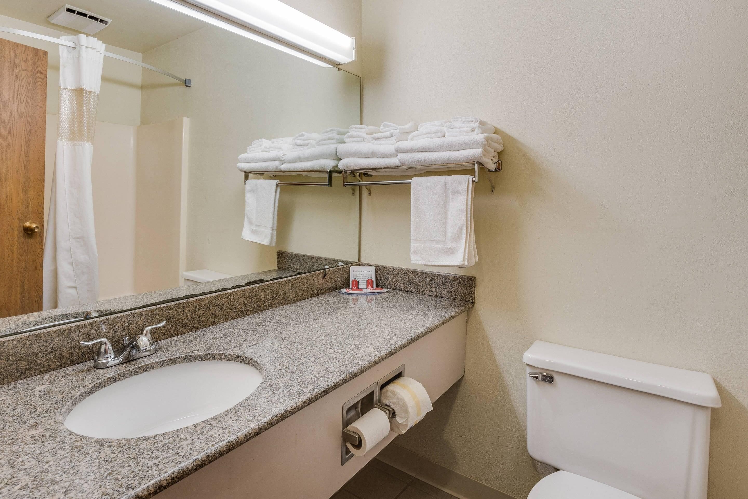 A bathroom with a granite countertop sink, a mirror, a towel rack with folded towels, a toilet, and a paper towel dispenser.