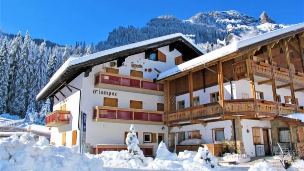 Un edificio de varios pisos con tejado cubierto de nieve y balcones, identificado como “Riampac”, rodeado por un paisaje nevado y montañas.