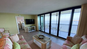 Un salón con un conjunto de sofá, una mesa de centro y un televisor. Hay un balcón con vista al océano.