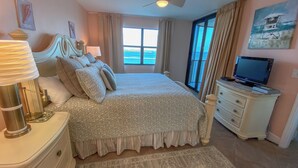 Un dormitorio con cama, televisor, cómoda, lámpara, ventilador de techo y un amplio ventanal con vistas al océano.