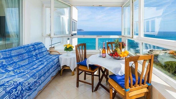 Un balcón con vista al océano, un sofá azul con estampado, una mesa de madera con sillas y una pequeña planta en maceta.